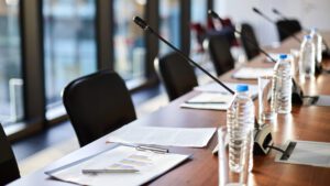 Business documents, plastic bottles of water, glasses and microphones along table and chairs in a board meeting