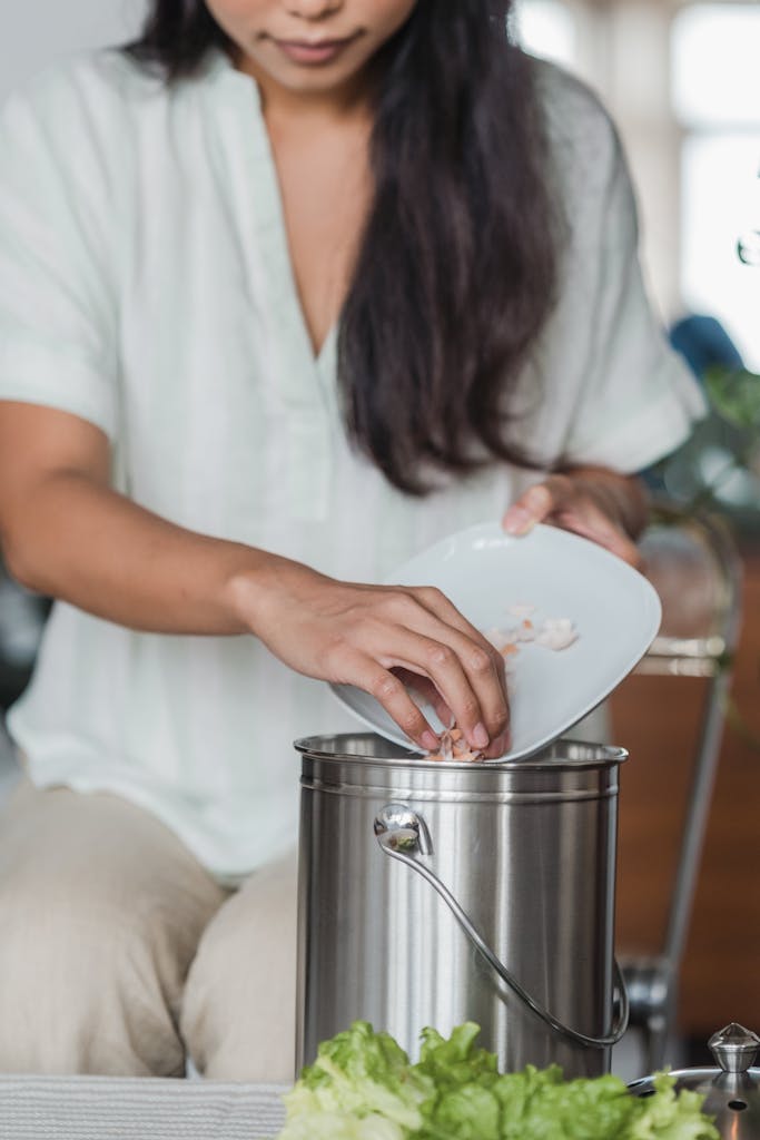 Close-up of a woman putting food scraps into a stainless steel compost bucket in the kitchen.