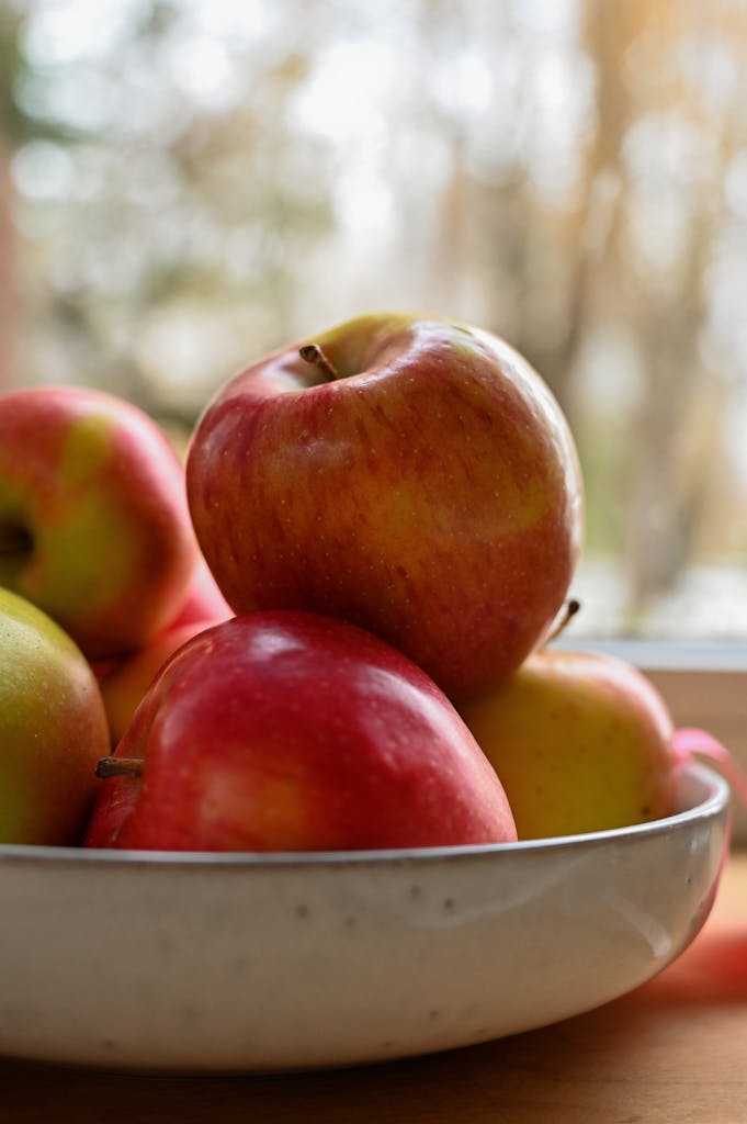 A stack of fresh red and green apples in a ceramic bowl by a window.