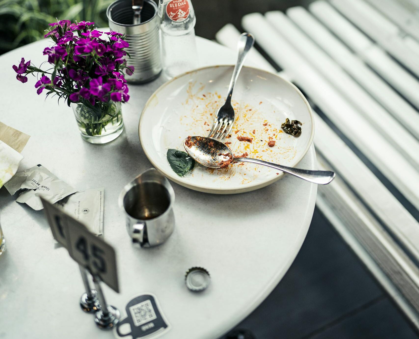 An indoor table setting with leftover food, cutlery, flowers, and a soda bottle.