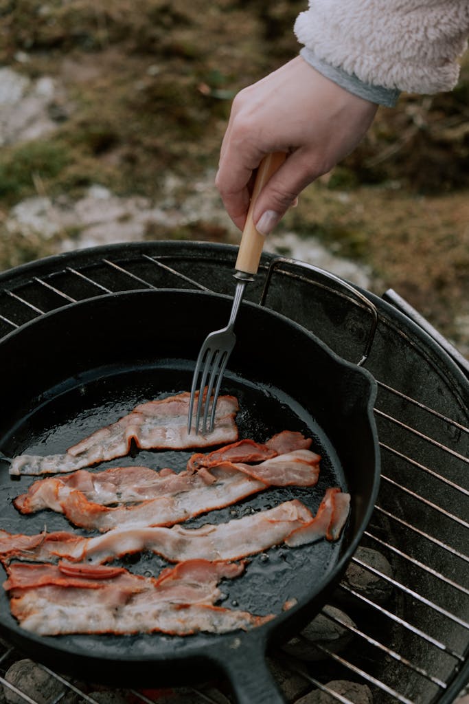 Close-up of bacon strips frying in a pan on a charcoal grill outdoors.